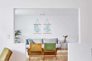 Ein schlicht eingerichtetes Wohnzimmer mit einem weißen Sofa, zwei Holzstühlen, einem kleinen Beistelltisch und zwei an der Wand befestigten Karten.