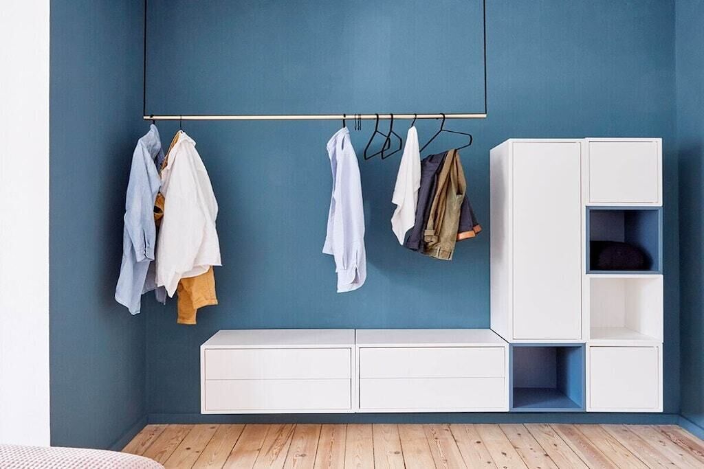 Ein Zimmer mit blauer Wand, Holzboden, weißem Kleiderschrank und Kleidung, die an einer Stange hängt.
