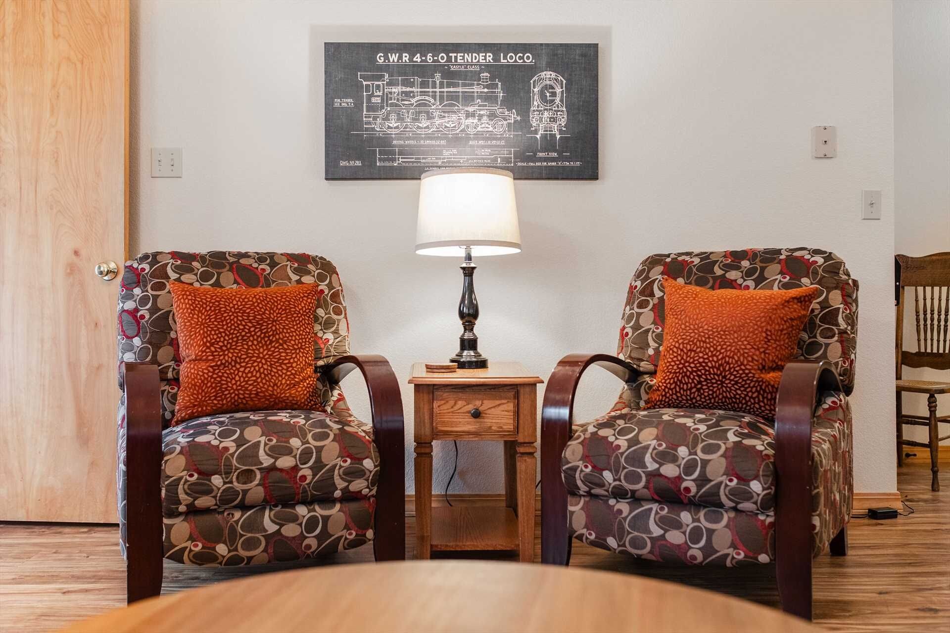 Een kamer met twee gestoffeerde fauteuils, een klein houten bijzettafeltje met een lamp, en een blauwdruk die aan de muur hangt.