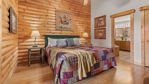 A log cabin-style bedroom with a bed, two bedside tables, a ceiling fan, and a bathroom visible through an open door.