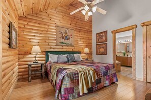 Ein schlafzimmer im Blockhausstil mit einem Bett, zwei Nachttischen, einem Deckenventilator und einem durch eine offene Tür einsehbaren Badezimmer.