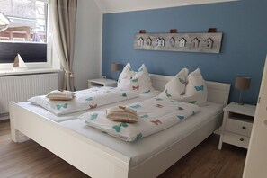 Een tweepersoonsbed met beddengoed met vlinderthema, een nachtkastje met een lamp, een wandplank met decoraties in de vorm van huisjes, en een raam met gordijnen.