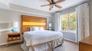 A hotel room with a bed, a ceiling fan, a nightstand with a lamp, a window with curtains, and a view of greenery outside. - Orlando | Orlando Resort Stay | Pool & Water Park | 2BR (Orlando)