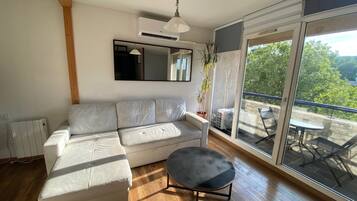 A modern living room with a white sofa, a small round table, a ceiling fan, and a large sliding glass door leading to an outdoor area with a table and chairs.