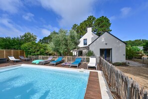 Ein Swimmingpool mit Liegestühlen, einer Holzterrasse und einem Gebäude mit Satteldach.