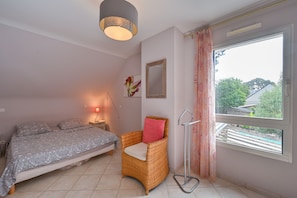 A bedroom with a bed, a chair, a bedside table with a lamp, a window with curtains, and a wall-mounted mirror. - Ker Augusta - Jolie maison 4 chambres avec piscine (Mesquer)