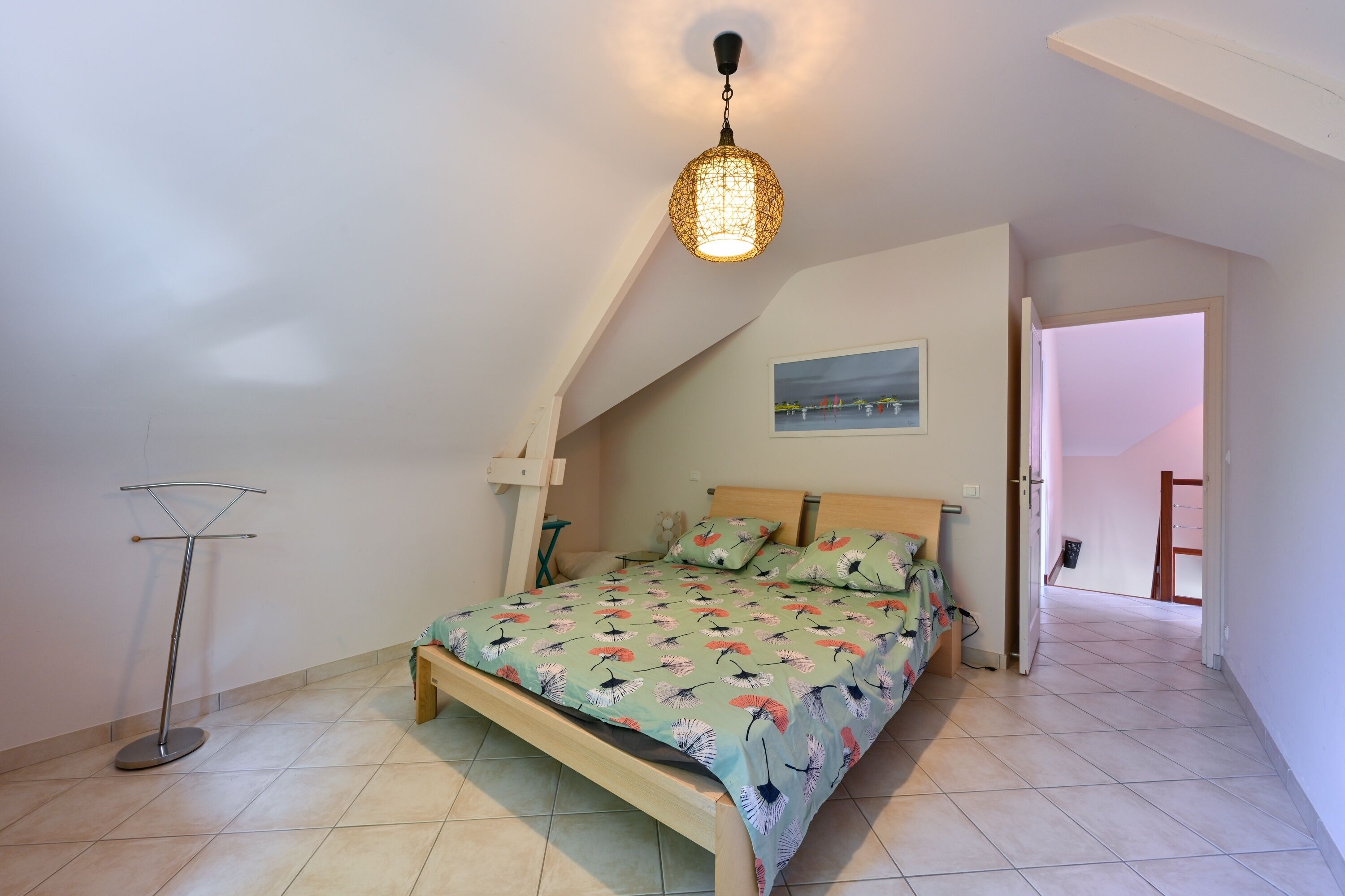 A bedroom with a bed, a hanging light fixture, a painting on the wall, and a view of the outside.