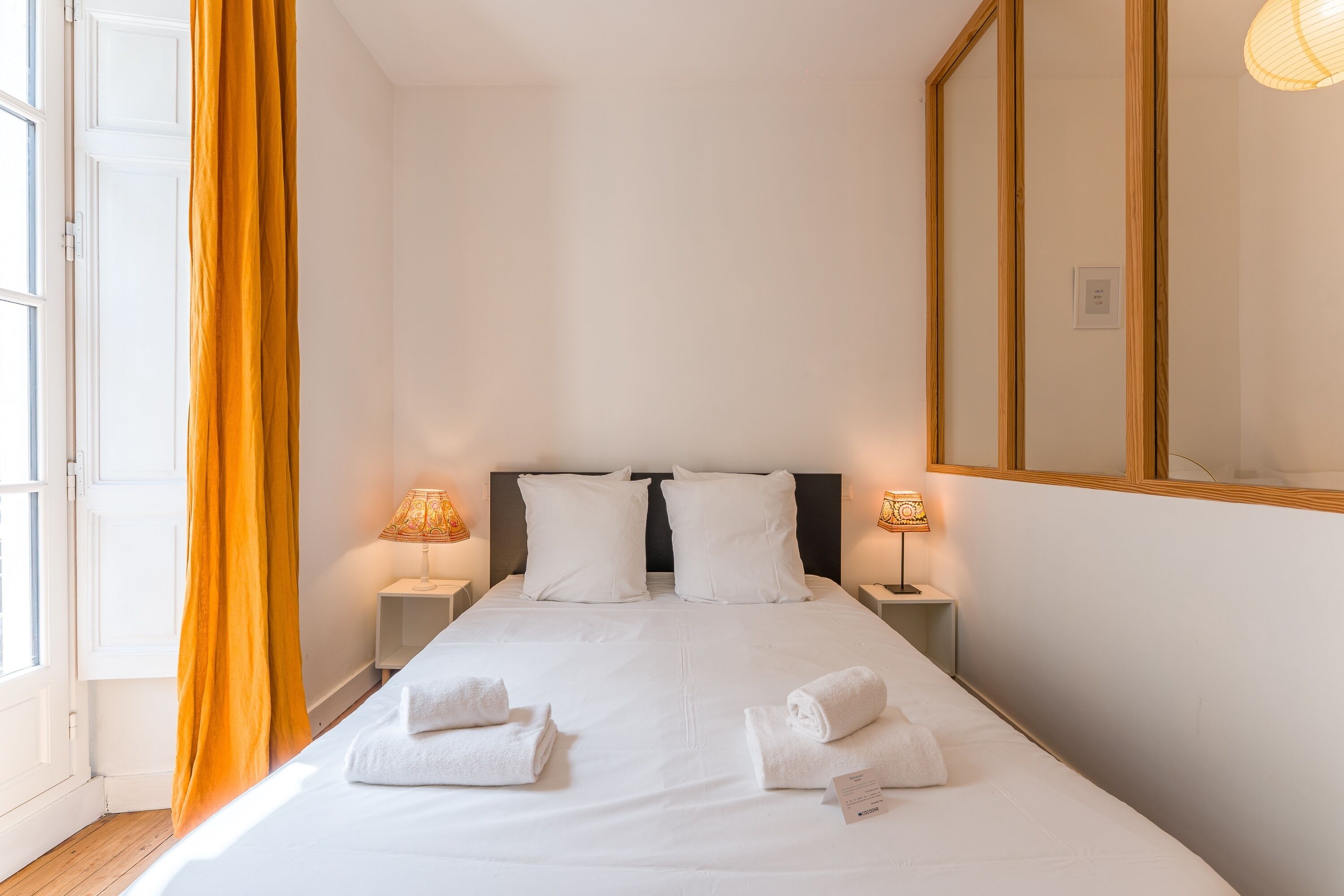Habitación de hotel con una cama, dos mesitas de noche, un espejo grande y una ventana con cortinas.