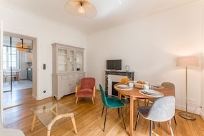 A dining area with a wooden table set for two, a red chair, a teal chair, and a grey chair. There is a white cabinet with glass doors, a television on a black stand, and a floor lamp. - Opaline - Bel appt 2 chambres au cœur de Nantes (Nantes)