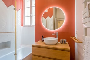 A bathroom with a white bathtub, a wooden vanity with a round mirror, and a small round container. - Opaline - Bel appt 2 chambres au cœur de Nantes (Nantes)