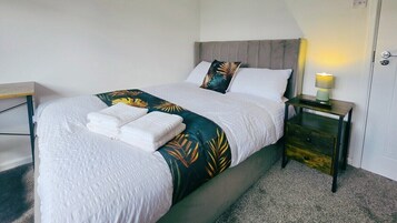 Una cama bien hecha con una colcha blanca y una manta con estampado de hojas tropicales, una mesita de noche con una lámpara y una puerta con pomo.