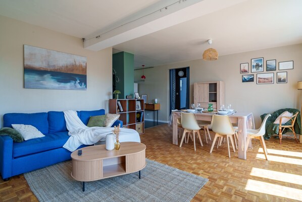 A modern living room with a blue sofa, a coffee table, and a dining area with a wooden table and chairs. - L’Écrin de Limoges - Grand studio avec parking (Limoges)