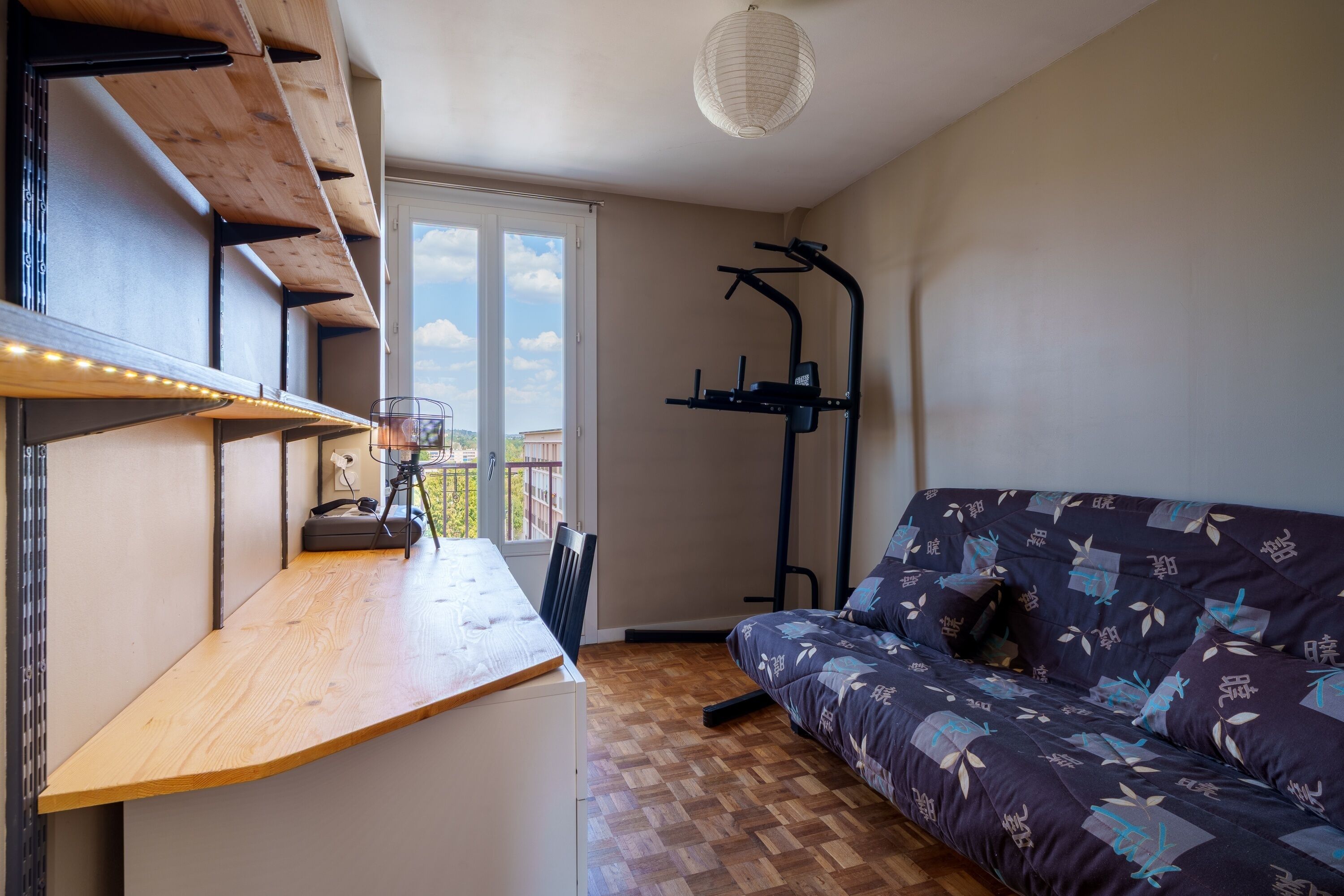 Una habitación con un escritorio de madera, un sofá con cojines estampados, una lámpara de techo y una ventana con vistas a edificios.