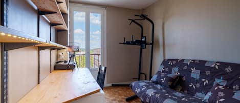 A room with a wooden desk, a sofa with patterned cushions, a ceiling light, and a window with a view of buildings.