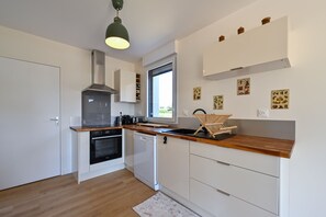 A modern kitchen with white cabinetry, a built-in oven, a dishwasher, and a window providing natural light. - Ty Liv - Maison familiale avec un beau jardin (Saint-Gildas-de-Rhuys)