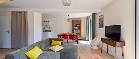 A modern living room with a grey sectional sofa, a wooden coffee table, a television on a wooden stand, and a dining area in the background.