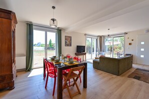 A spacious dining area with a wooden table and red chairs, a television, and a sofa in a bright room with large windows. - Ty Liv - Maison familiale avec un beau jardin (Saint-Gildas-de-Rhuys)