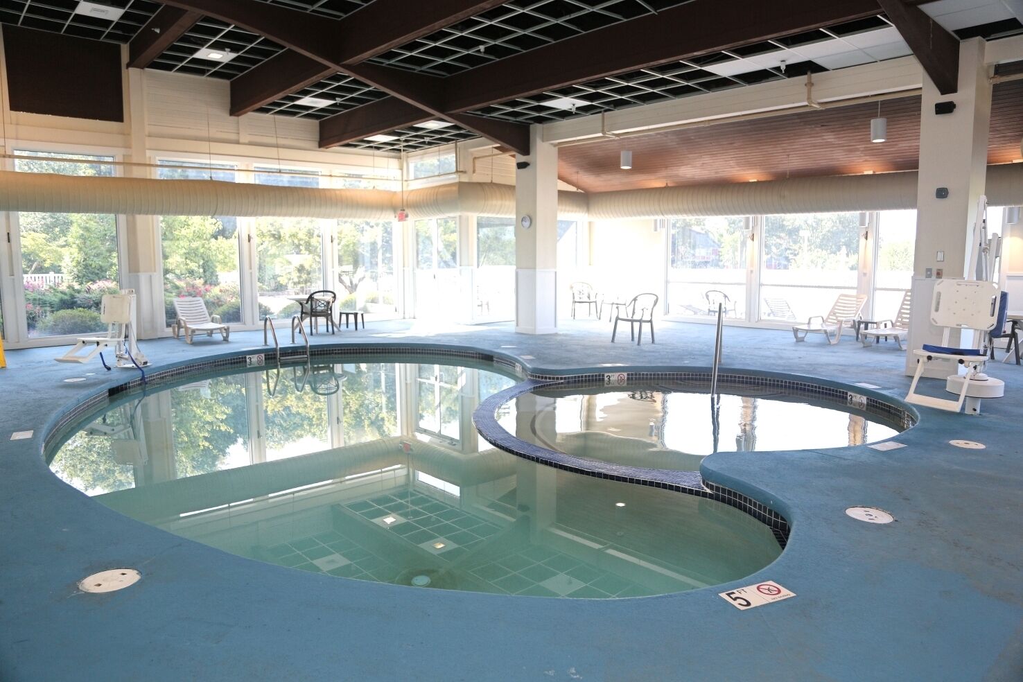 An indoor swimming pool with a curved design, surrounded by lounge chairs and tables, large windows, and a high ceiling with exposed beams.