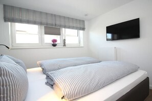 A bedroom with two beds, a window with blinds, a flat-screen TV mounted on the wall, and a bedside table with a vase. - Fischerweg 15i Ground Floor (Sylt \/ OT Westerland)