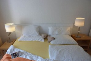 Una habitación de hotel con una cama, dos lámparas de noche y un osito de peluche sobre las almohadas.