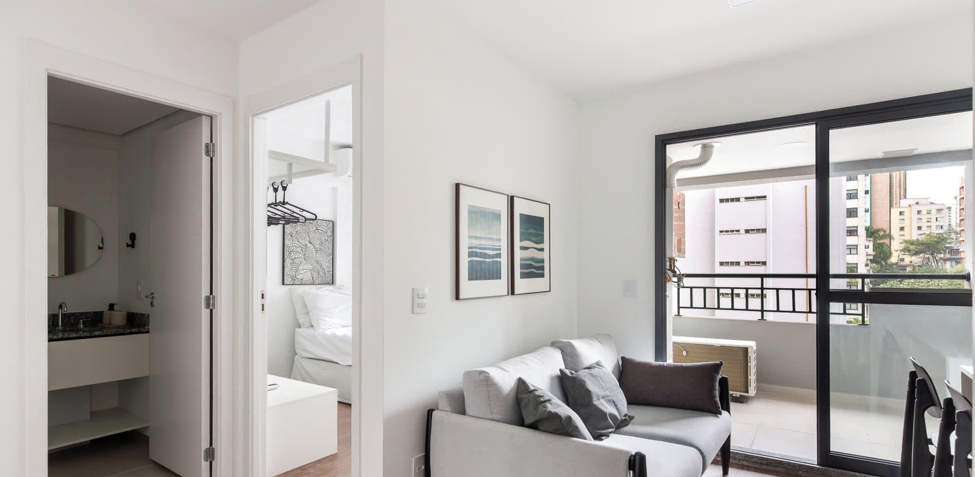 A modern living room with a sofa, a balcony with a view of buildings, and a door leading to a bathroom.