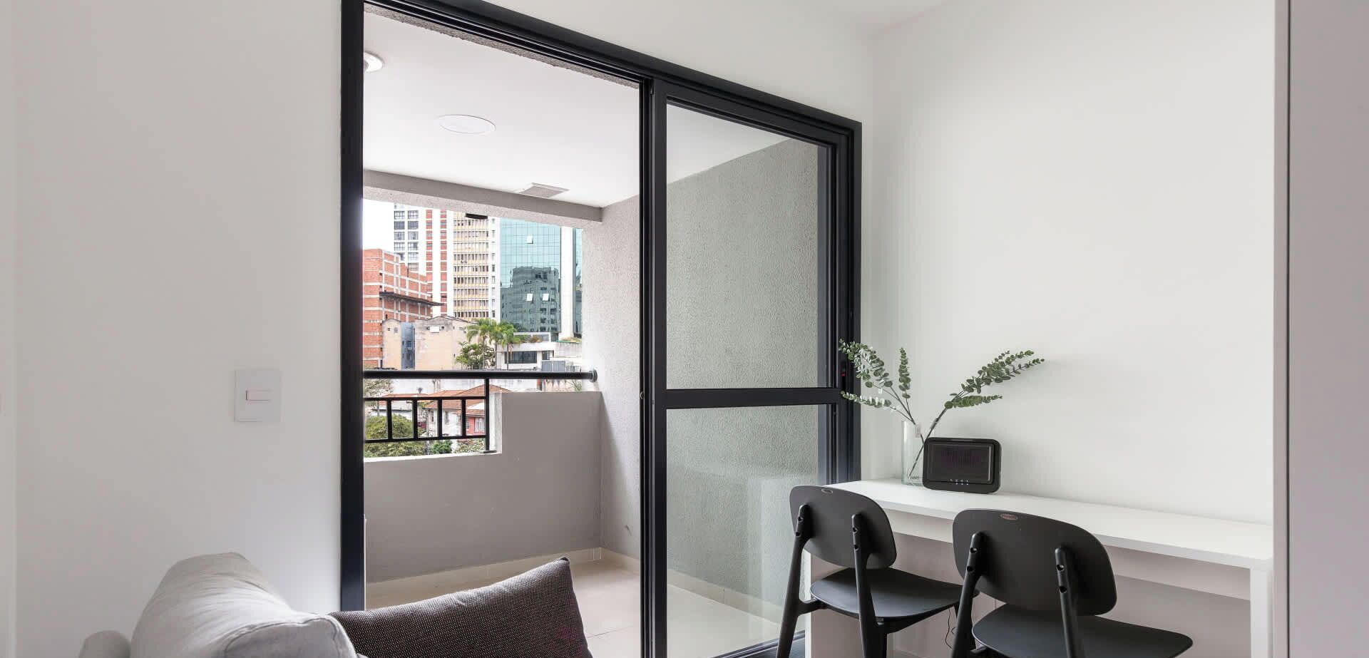 A modern apartment with a balcony, a white sofa, two black chairs, a small table, and a vase with plants.