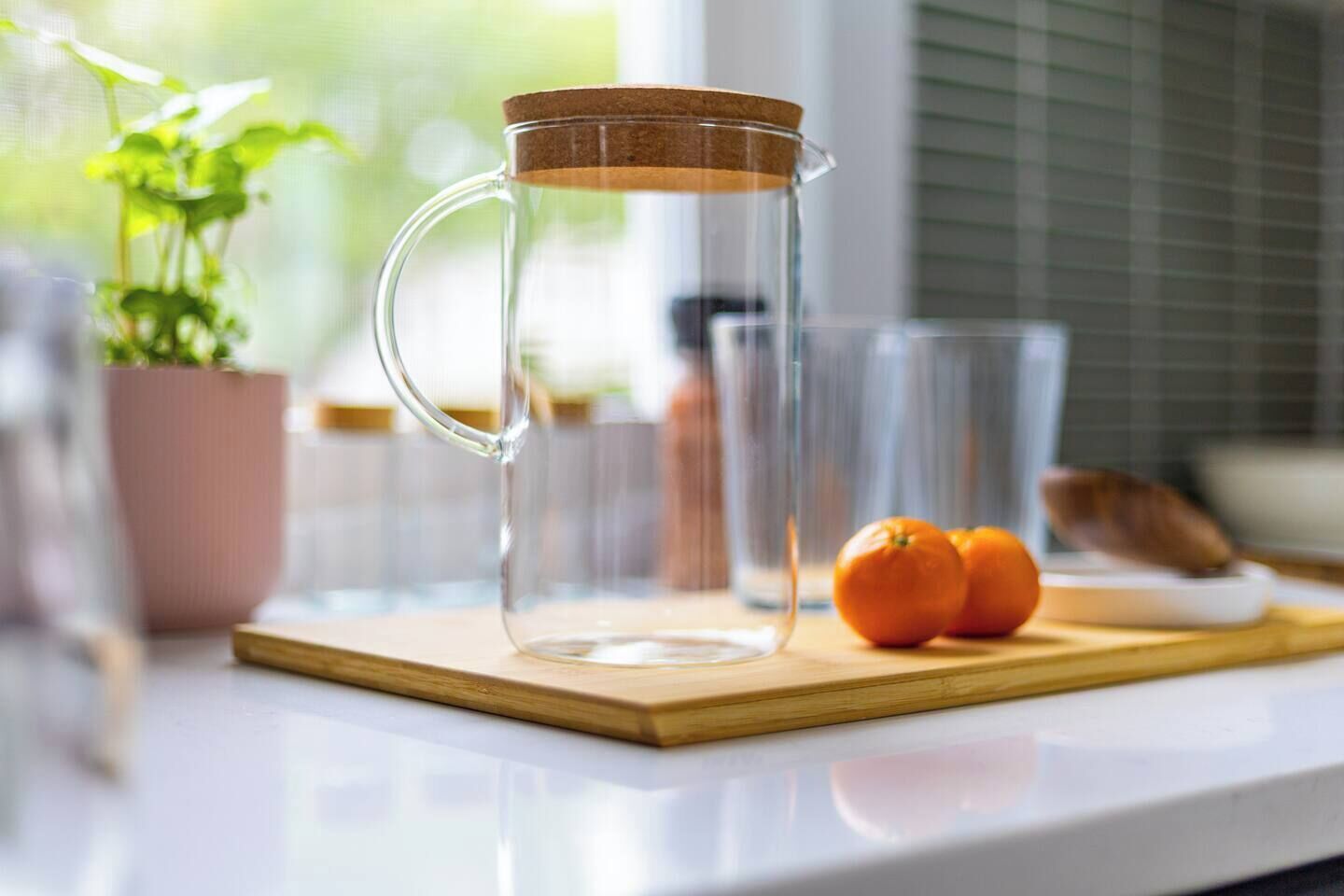 Um jarro de vidro transparente com rolha de cortiça sobre uma tábua de madeira, um copo de água e uma planta em vaso ao fundo.