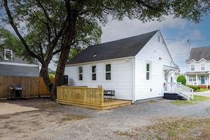 Ein eingeschossiges weißes Haus mit Veranda, einer Holzterrasse und einem großen Baum davor.