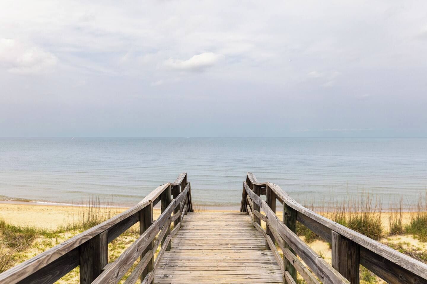 Ein Holzsteg führt zu einem Strand mit ruhiger See und bewölktem Himmel.