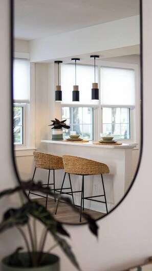 Cozinha com bancada branca, dois banquetes de vime, luminárias pendentes e janela com vista.