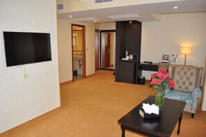 A hotel room with a flat-screen TV mounted on the wall, a small table with a vase of flowers, a sofa with a patterned cushion, and a desk with a lamp.