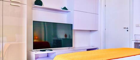 A modern hotel room with a large flat-screen TV, white furniture, and a bed with a yellow bedspread.