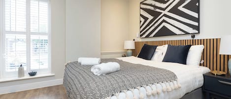 A hotel room with a large bed, a bedside table with a lamp, a wall-mounted artwork, and a window with blinds.