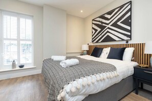 A hotel room with a large bed, a bedside table with a lamp, a wall-mounted artwork, and a window with blinds. - Stylish Modern Studio with Double Bed (Southampton)