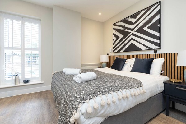 A hotel room with a large bed, a bedside table with a lamp, a wall-mounted artwork, and a window with blinds. - Stylish Modern Studio with Double Bed (Southampton)