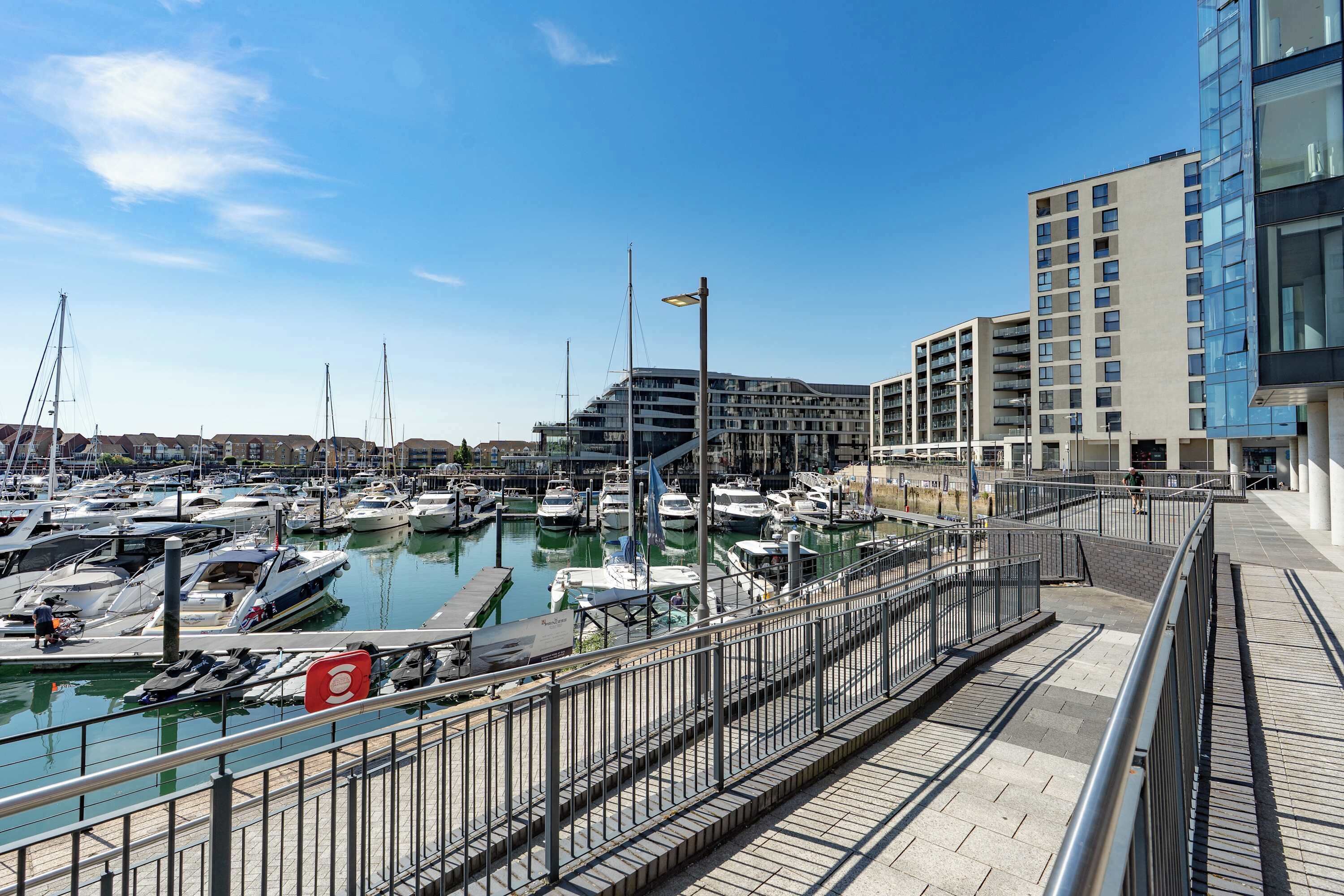 A marina with boats docked, a pedestrian walkway, and modern buildings in the background.
