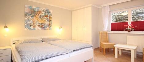 Un dormitorio con cama, una silla, una mesita, una ventana con cortinas y un mapa colgado en la pared.
