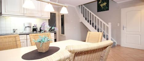 Un comedor con sillas de mimbre y una mesa, una cocina con armarios blancos y una escalera que sube a otro nivel.