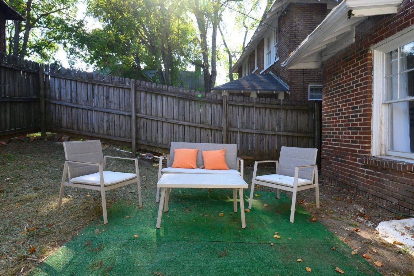 Un patio trasero con cerca de madera, una casa de ladrillo y mobiliario exterior, incluyendo dos sillas y una mesa.