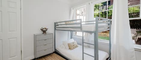 A bunk bed with a view of the outdoors, a small dresser with drawers, and a window with curtains.