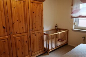 A room with a wooden wardrobe, a crib with a baby, and a window with a curtain. - Holiday home with 3 bedrooms (Wangerland)