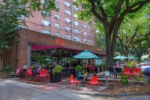 An outdoor seating area with tables and chairs, a brick building with a neon sign, and trees in the background. - Historic Highland Park Gem (Birmingham)