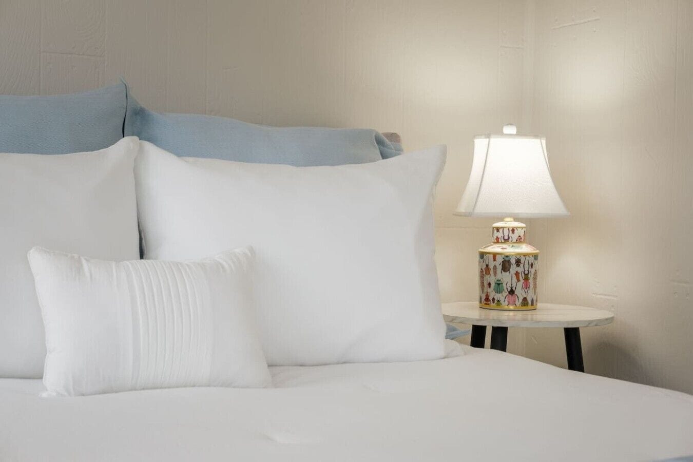 A bed with white bedding and pillows, a round table with a lamp, and a wall with a textured pattern.