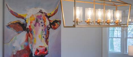 Una habitación con un cuadro abstracto de una vaca, una lámpara de araña geométrica y un ventanal con vistas.