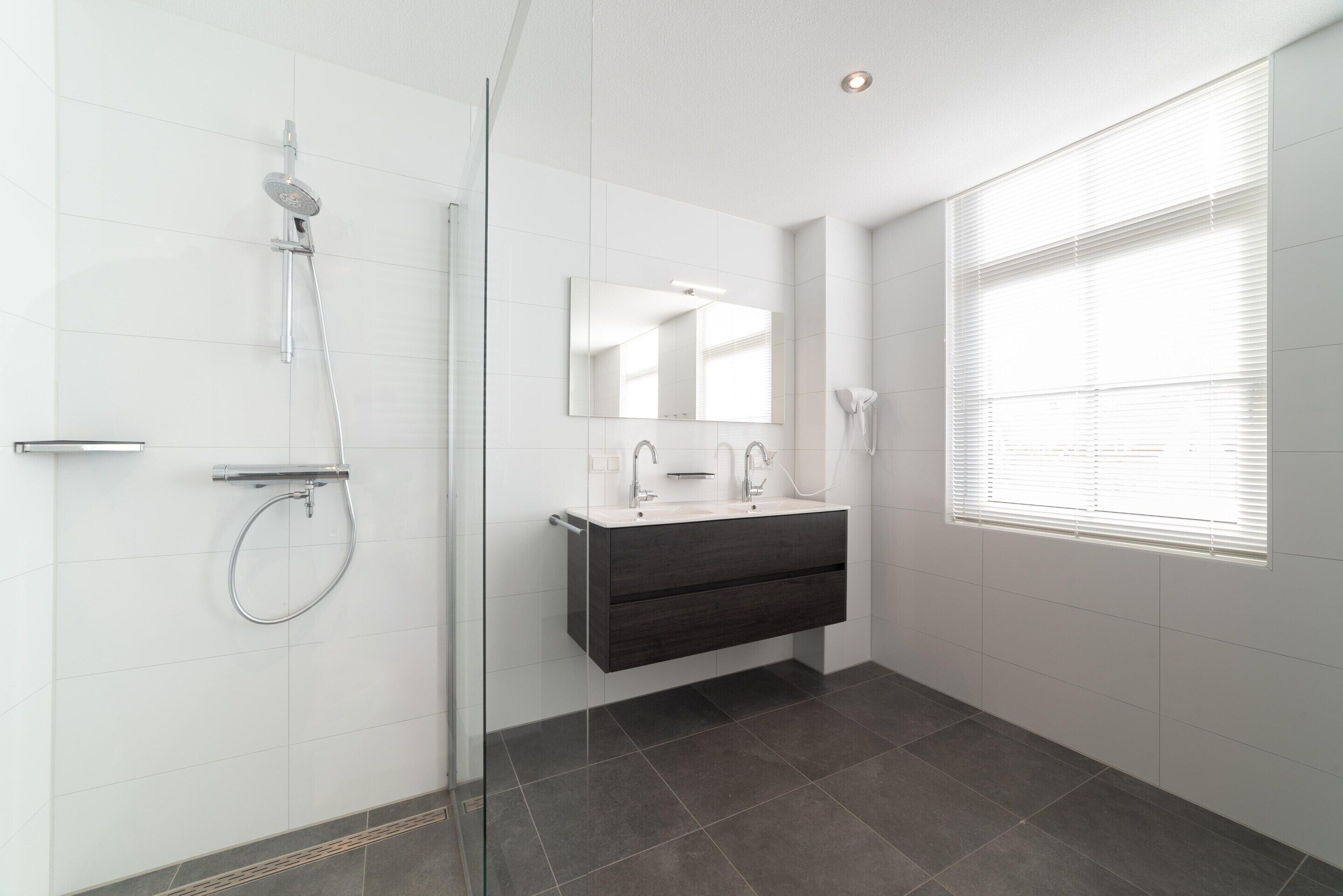 A modern bathroom with a glass shower enclosure, a dark vanity, a large mirror, and a window providing natural light.
