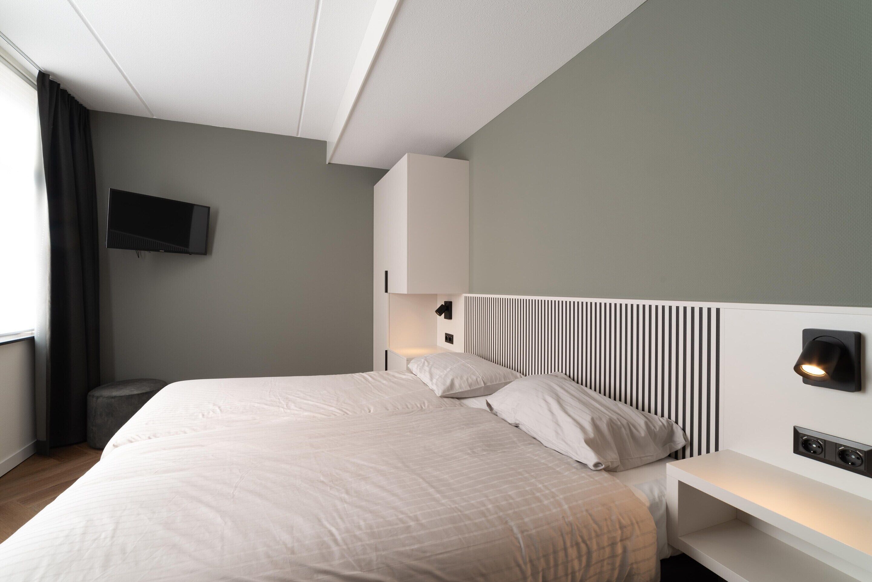 A modern bedroom with a large bed, a flat-screen TV mounted on the wall, a bedside table with a lamp, and a built-in headboard with vertical slats.