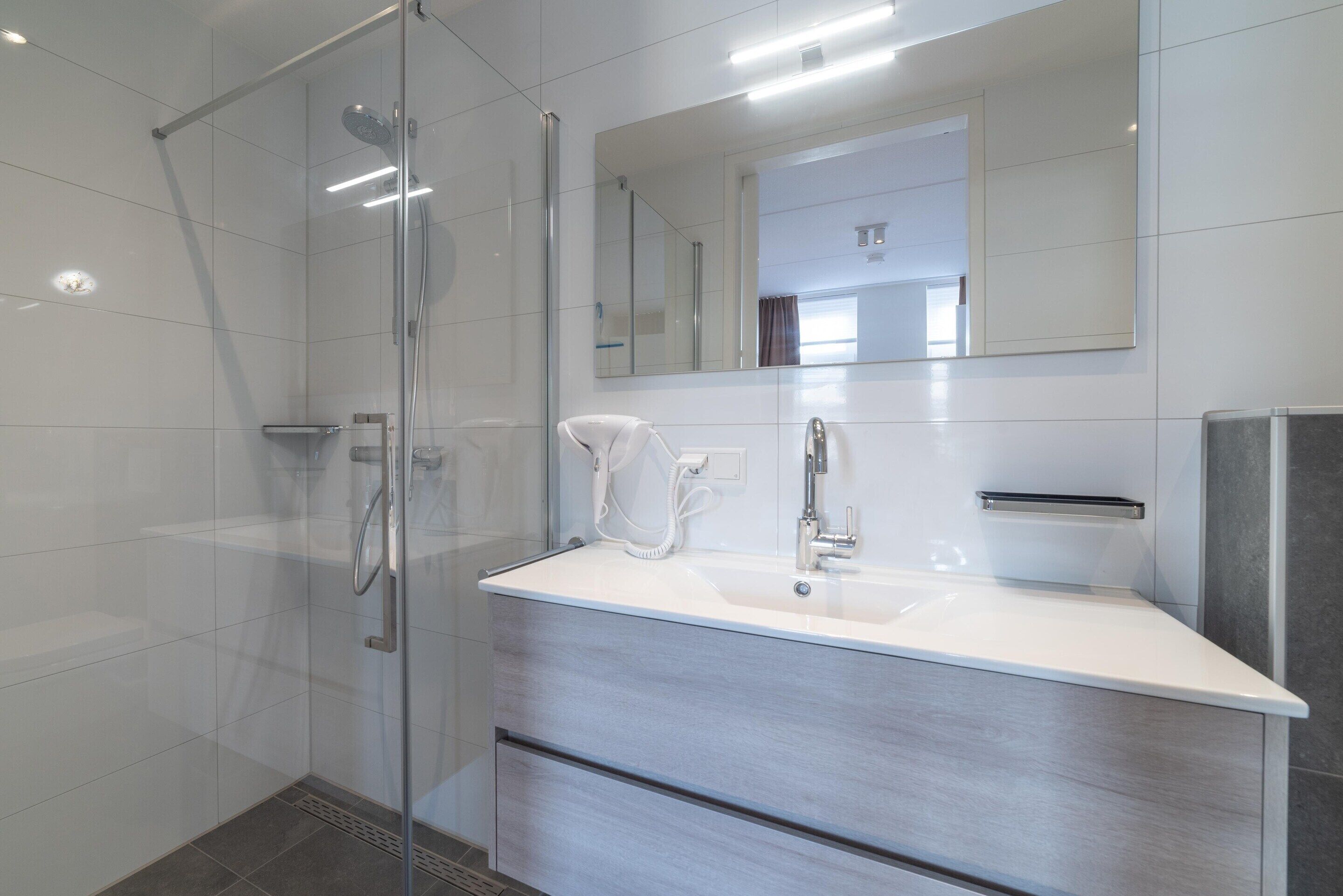 Une salle de bain moderne avec une douche à l’italienne en verre, un lavabo blanc et un grand miroir.