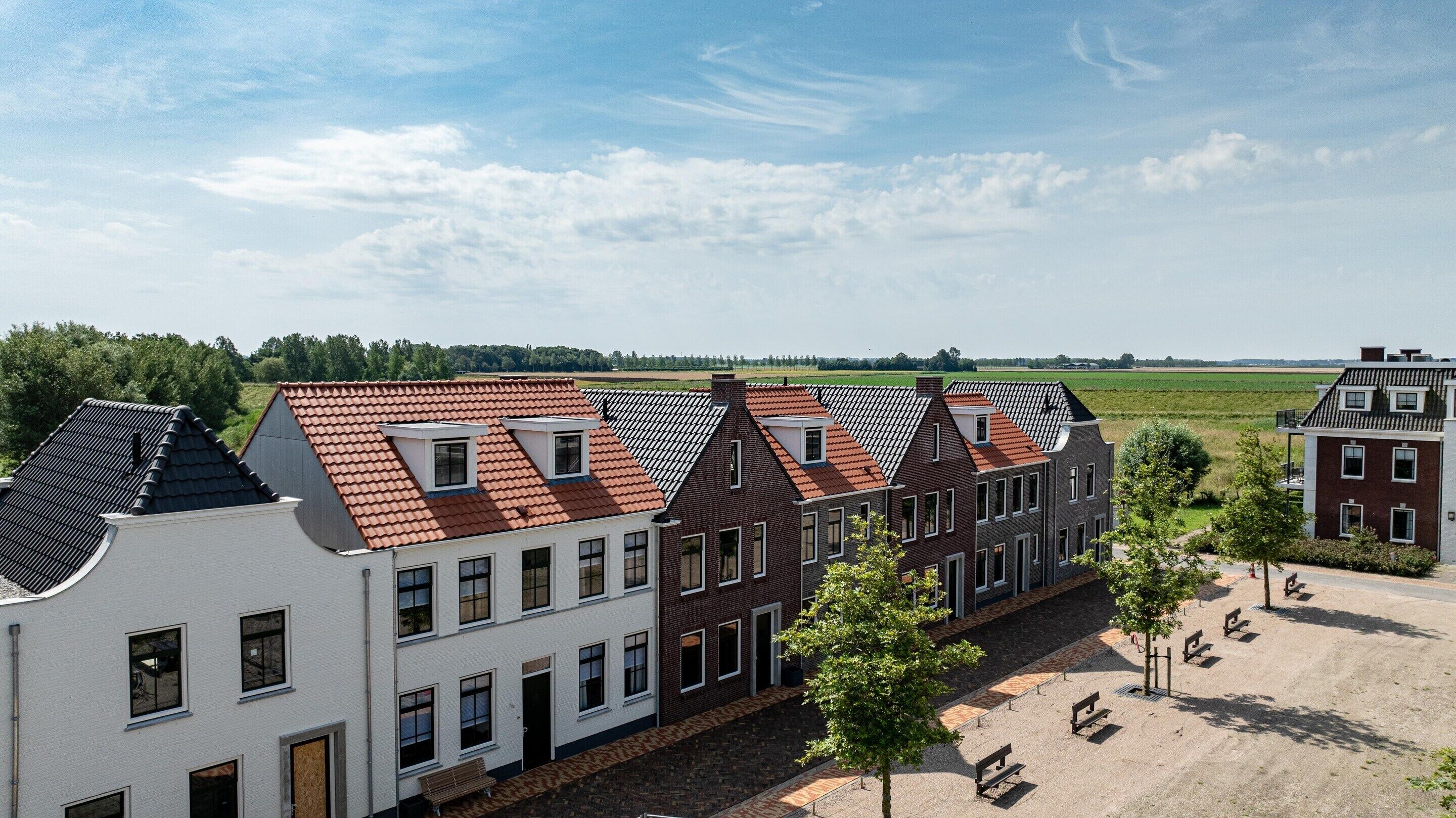 A row of traditional European-style houses with red-tiled roofs and white walls, set in a rural area with open fields and a clear sky.