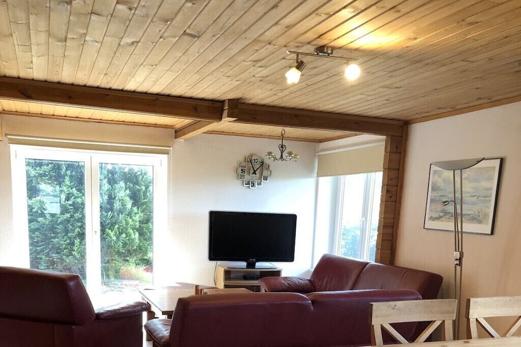 A living room with a wooden ceiling, a television, a sofa, and a window with a view of greenery.