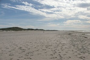 Ein weiter Sandstrand mit Fußabdrücken, sanften Hügeln in der Ferne und einem teils bewölkten Himmel.